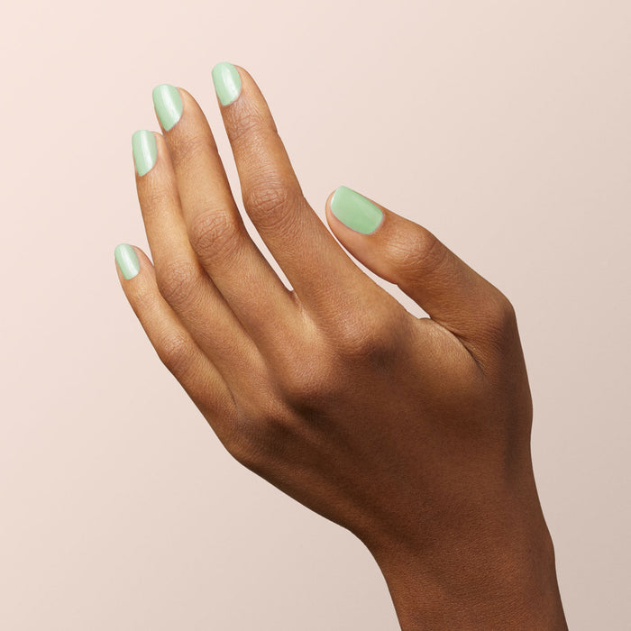 A hand with medium brown skin is shown against a light beige background. The neatly manicured fingernails are painted with Manucurist Mint, a pastel green, glossy Green Flash gel polish.