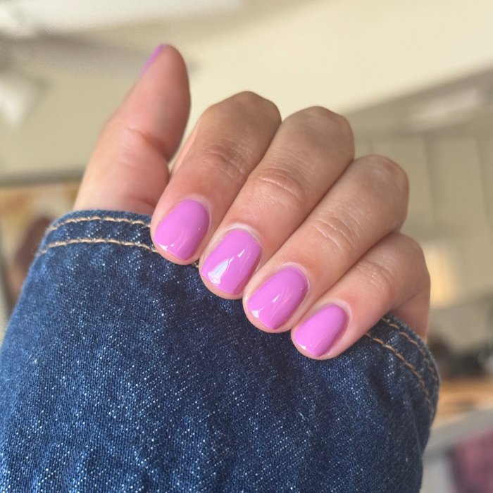 Hand with short rounded nails painted in bright floral purple pink using Petal - Gel against casual denim background