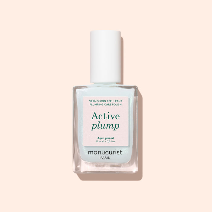 A bottle of Manucurist Active™ Plump Aqua Glazed plumping care polish with a white cap is displayed against a pale pink background. This volumizing nail care formula, featuring collagen nail polish, presents its label text on the front of the clear bottle.