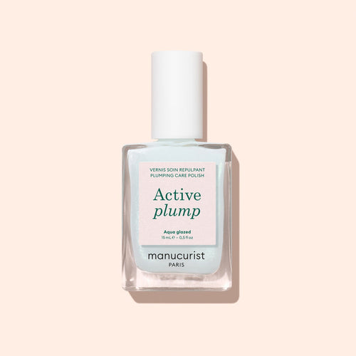 A bottle of Manucurist Active™ Plump Aqua Glazed plumping care polish with a white cap is displayed against a pale pink background. This volumizing nail care formula, featuring collagen nail polish, presents its label text on the front of the clear bottle.