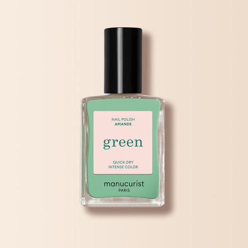 A bottle of Manucurist Amande nail polish from the Green™ range, featuring a mint green shade and black cap, labeled "Quick Dry Intense Color," is displayed on a light beige background.