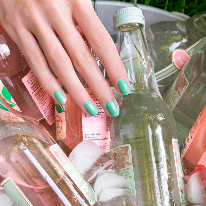 A hand with Manucurist’s Amande green nail polish rests on ice-cold beverage bottles, some featuring pink labels, in a metal tub filled with ice.