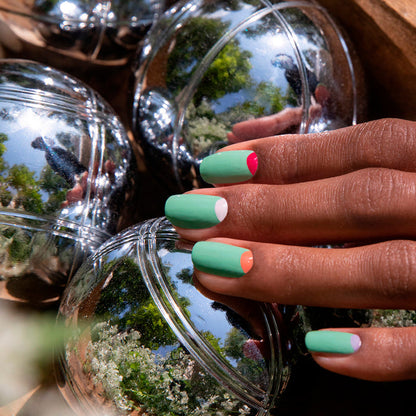 A hand with colorful, half-moon manicured nails—including Manucurist’s Amande shade—rests on shiny metallic balls reflecting trees and sky.