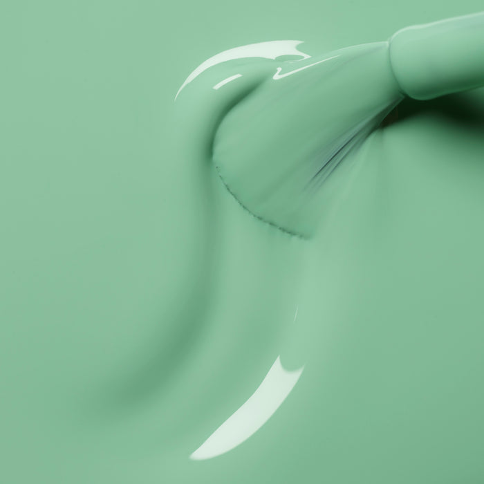 A close-up of a brush applying Manucurist’s Amande glossy mint green polish in smooth strokes, highlighting visible texture and light reflections on the wet surface.
