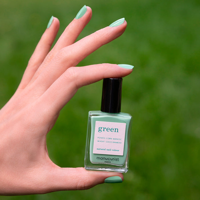 A hand with mint green painted nails holds a bottle of Amande nail polish by Manucurist against a blurred green background.