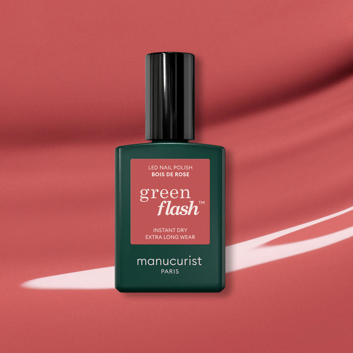 A bottle of Manucurist Bois de Rose gel polish stands upright on a pink background with a white brushstroke. The dark green bottle features a black cap, pink label, and displays its elegant rosewood pink shade.