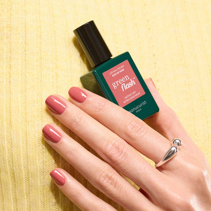 A hand with neatly manicured pink nails holds a Manucurist Bois de Rose Green Flash gel polish bottle against a yellow textured background, sporting a silver ring with two round ends.