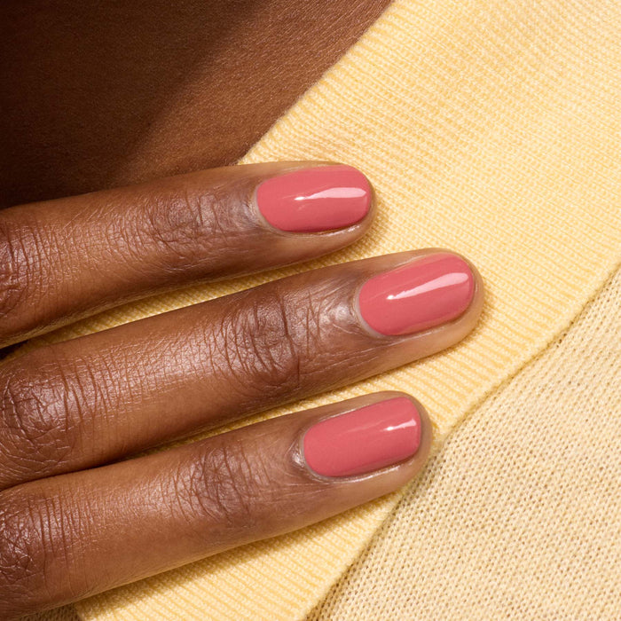 A close-up of a hand with short, neatly manicured nails painted in Manucurist's glossy Bois de Rose shade, resting on yellow knit fabric.
