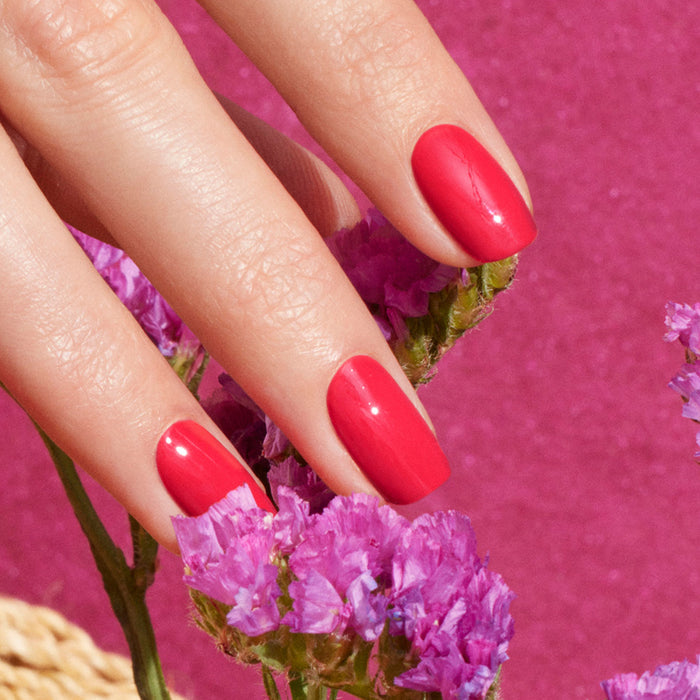 A close-up of a hand with Manucurist’s Bougainvillea, a shiny reddish pink nail polish, holding small bougainvillea flowers against a vibrant pink background.