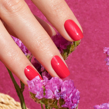 A close-up of a hand with Manucurist’s Bougainvillea, a shiny reddish pink nail polish, holding small bougainvillea flowers against a vibrant pink background.