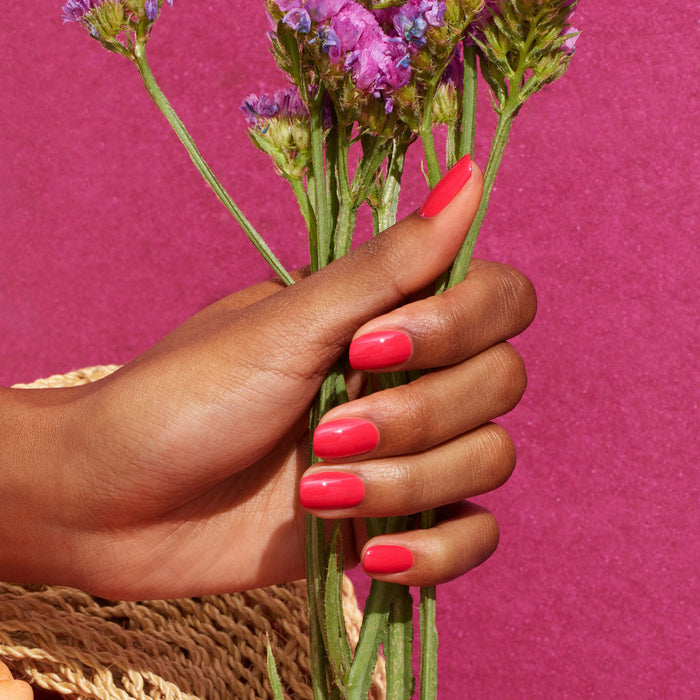 A hand with glossy, reddish pink Manucurist Bougainvillea nail polish holds a small bouquet of bougainvillea against a textured pink background.