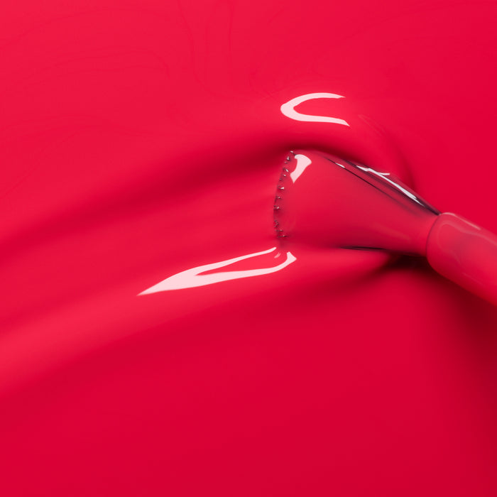 A close-up of a makeup brush spreading Manucurist Bougainvillea, a bright reddish-pink nail polish with glossy reflections on the smooth surface.
