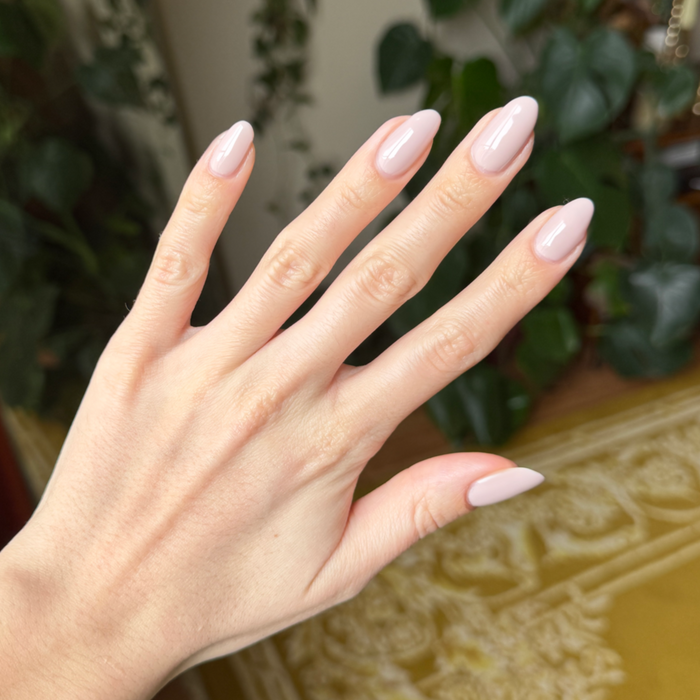 Hand with long almond-shaped nails painted in CDJ - Gel creamy neutral gel polish against blurred indoor background