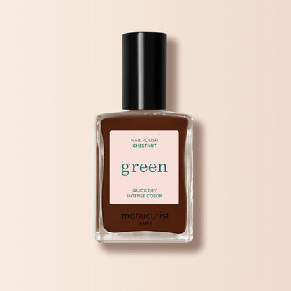 A bottle of Manucurist Chestnut nail polish stands upright on a beige background. The label reads "green, quick dry intense color," and the warm brown shade features a rich, hazelnut tone.