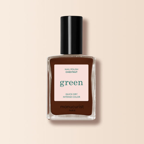 A bottle of Manucurist Chestnut nail polish stands upright on a beige background. The label reads "green, quick dry intense color," and the warm brown shade features a rich, hazelnut tone.