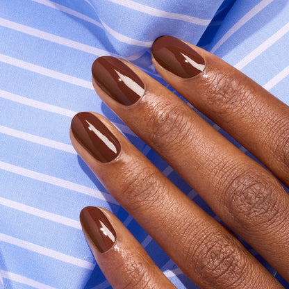 A hand with neatly manicured nails painted in glossy Chestnut nail polish from Manucurist rests on light blue fabric with white stripes.