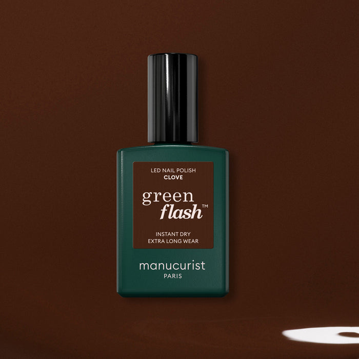 A bottle of Manucurist Clove gel polish, a rich chocolate brown shade, is shown against a brown background with a small light spot near the lower right corner.