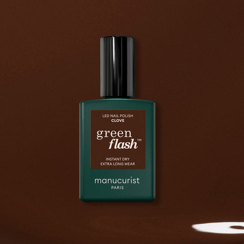 A bottle of Manucurist Clove gel polish, a rich chocolate brown shade, is shown against a brown background with a small light spot near the lower right corner.