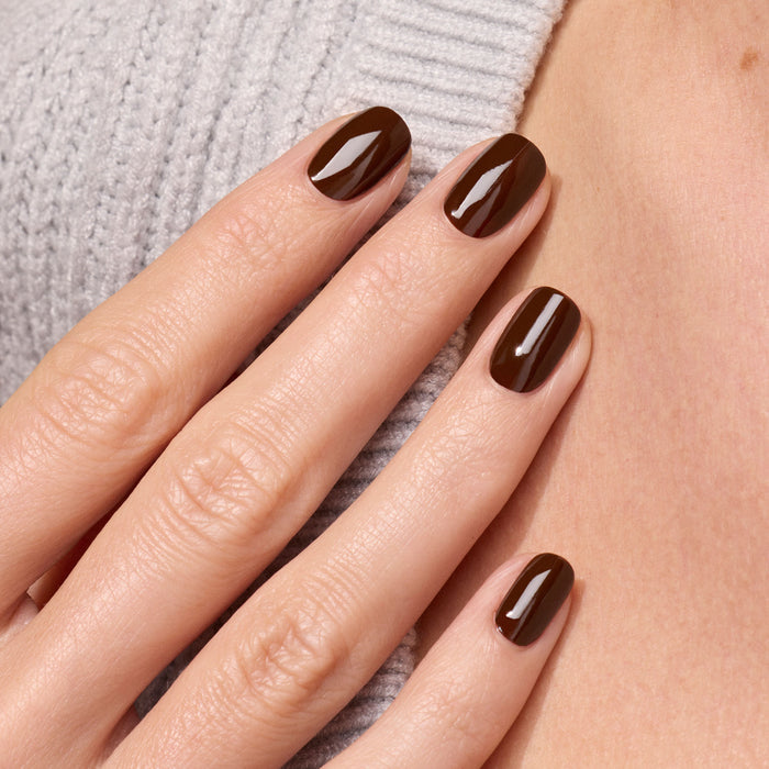 A close-up of neatly manicured nails painted in Manucurist’s Clove, a glossy chocolate brown gel polish, gently touching the neck. The person is wearing a light gray ribbed sweater.