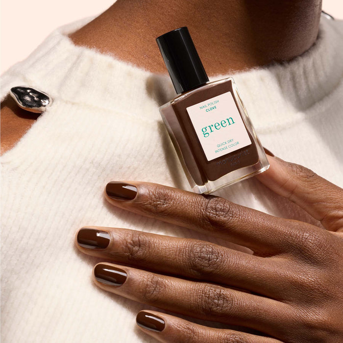 A hand with neatly manicured Clove brown nails from Manucurist holds a bottle labeled "green," set against a cream-colored sweater with button details.