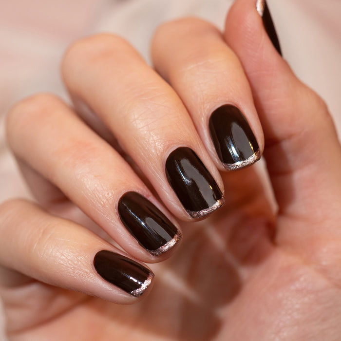 A hand with neatly manicured nails painted in Clove by Manucurist, a chocolate brown shade, each nail accented with a thin metallic gold line at the tips for a modern, elegant style.