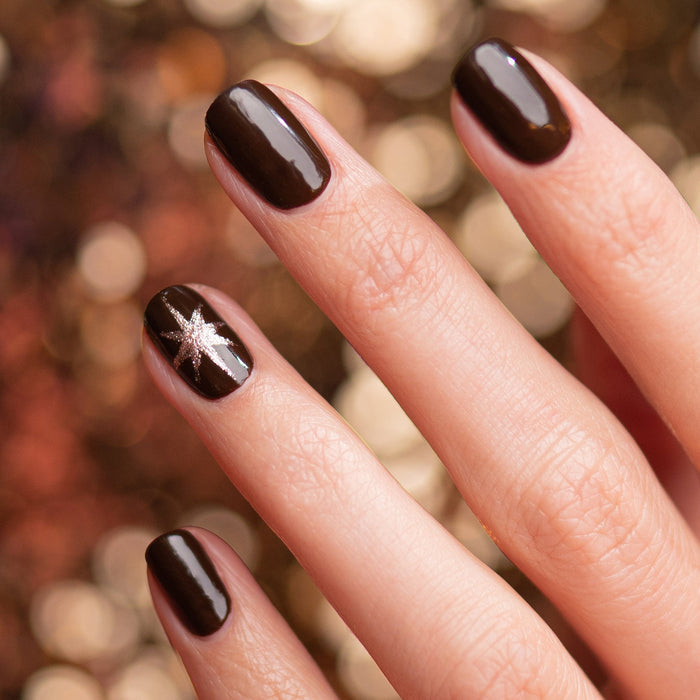 A hand with neatly manicured nails painted in Manucurist’s Clove, a glossy chocolate brown gel polish. The ring fingernail has a silver star design. The background is softly blurred with warm golden bokeh lights.