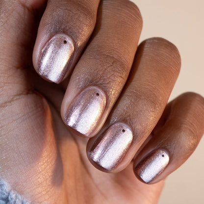 A hand with Manucurist's Cosmic Rose polish—metallic rose gold and iridescent pink shades, each nail topped with a black dot near the base—displayed against a neutral background.
