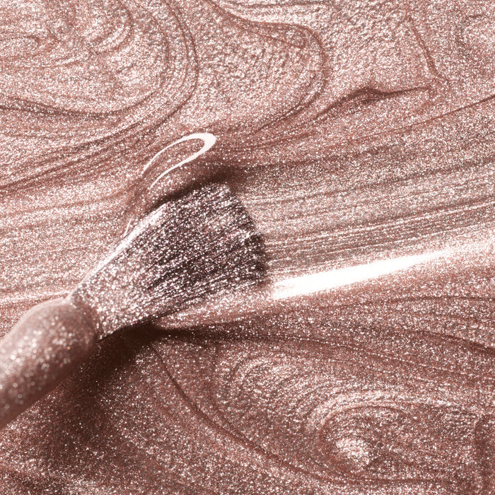 A close-up of a makeup brush spreading Manucurist's Cosmic Rose nail polish—a shimmery, iridescent pink with rose gold tones, swirling patterns, and a glossy finish.