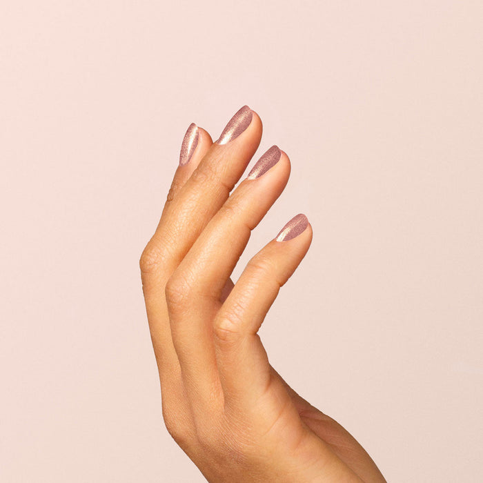 A hand with medium skin tone is shown against a light beige background, nails manicured and painted with Manucurist’s Cosmic Rose for an iridescent, shimmery mauve-pink metallic finish.