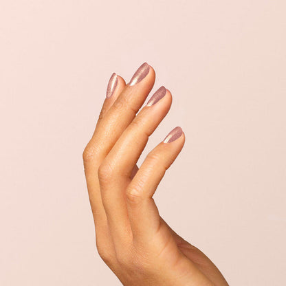 A hand with medium skin tone is shown against a light beige background, nails manicured and painted with Manucurist’s Cosmic Rose for an iridescent, shimmery mauve-pink metallic finish.