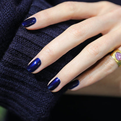 A close-up of a hand with Manucurist Dark Night gel polish, resting on a navy blue knitted sleeve. The ring finger features a gold ring with a pink stone.