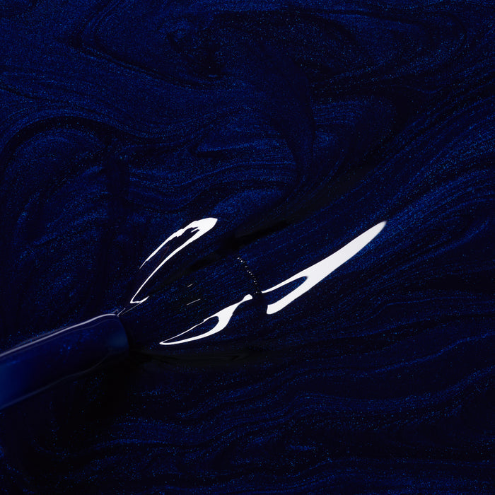 A close-up of a brush applying Manucurist’s Dark Night gel polish, revealing swirling midnight blue patterns and glossy, reflective highlights on the nail surface.