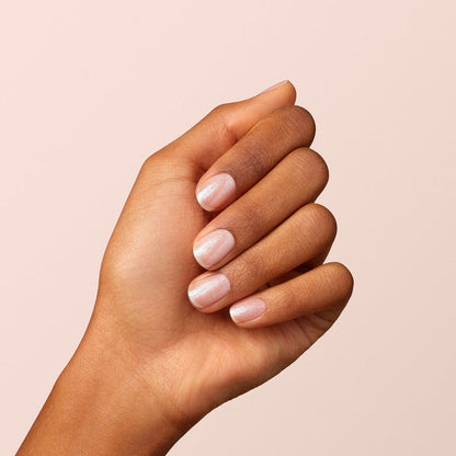 A hand with medium skin tone shows neatly manicured nails painted with Manucurist's Diamant, a shimmery light pink and silver nail polish, against a plain light beige background.