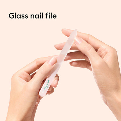 A person shapes their nails with a Manucurist-us Glass nail accessory trio, featuring a clear glass file as "Glass nail file" appears at the top left on a pale background. A glass cuticle pusher complements the manicure routine.