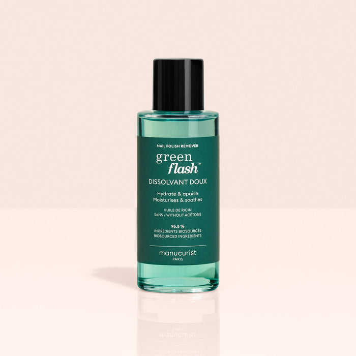 A bottle of Manucurist-us Green Flash™ Remover  Clips Duo, featuring green liquid and a black cap, rests on a light pink surface. The label notes its hydrating, soothing, acetone-free formula.