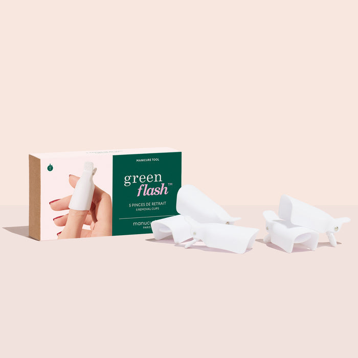 A box labeled Green Flash™ Remover  Clips Duo by Manucurist-us is shown with a hand wearing a white nail cap. Five white reusable clips for gel polish removal are displayed on a light surface beside the box.