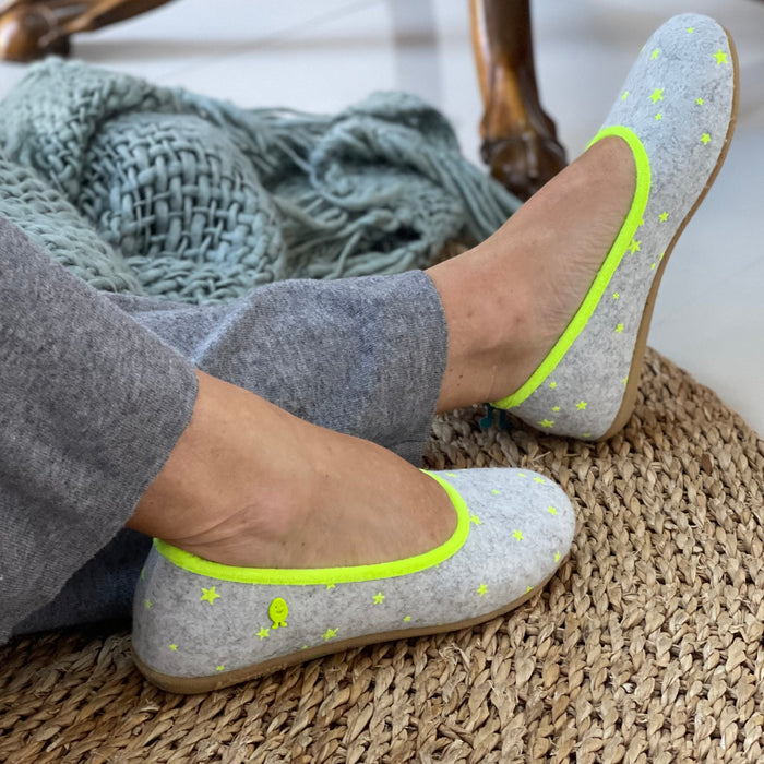 A pair of gray Hot Potatoes Mace slippers with yellow piping and star patterns being worn on a person's feet, with a carpeted floor and a cozy room setting in the background. Comfortable Spanish slippers/house shoe.