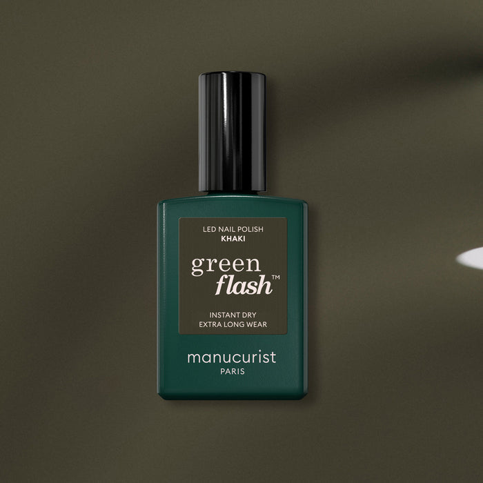 A bottle of Manucurist Khaki gel polish with gray undertones, dark green label, and black cap rests on an olive green surface.