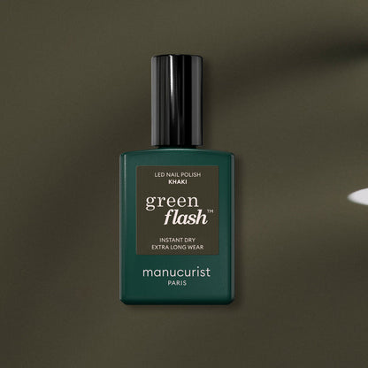 A bottle of Manucurist Khaki gel polish with gray undertones, dark green label, and black cap rests on an olive green surface.