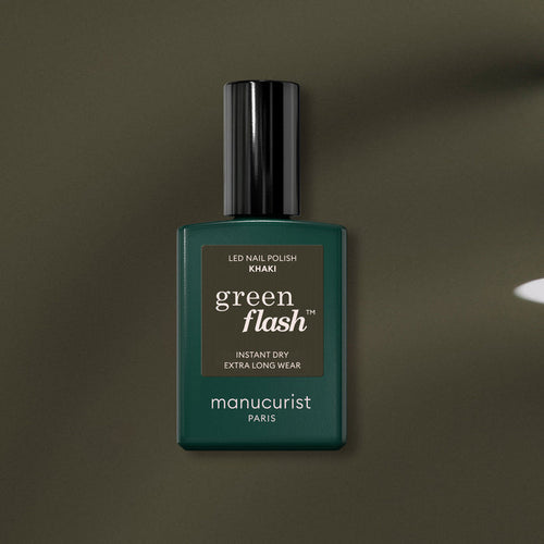 A bottle of Manucurist Khaki gel polish with gray undertones, dark green label, and black cap rests on an olive green surface.