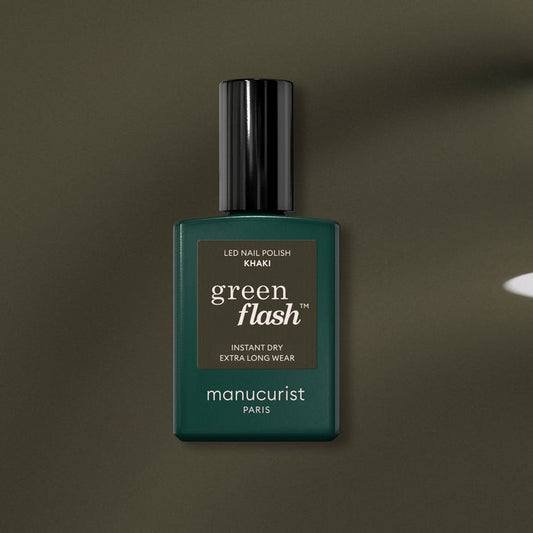 A bottle of Manucurist Khaki gel polish with gray undertones, dark green label, and black cap rests on an olive green surface.