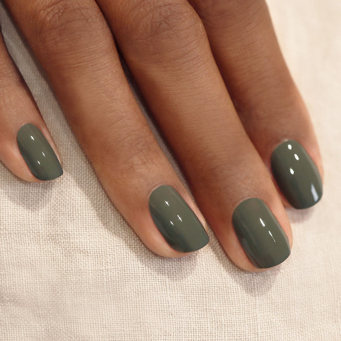 A hand with short, neatly manicured nails painted in Manucurist's Khaki—a glossy olive green shade with subtle grey undertones—rests on a light beige fabric background.