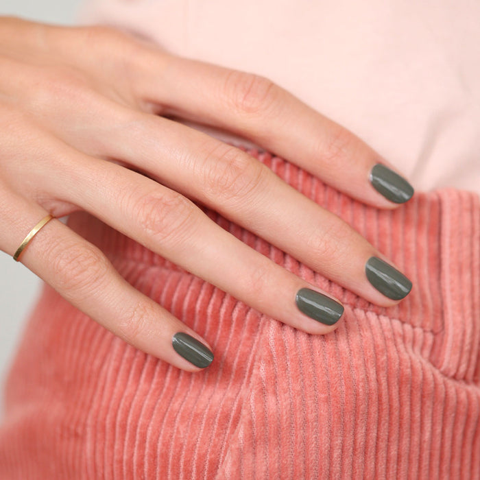 A hand with short, neat nails painted in Manucurist's Khaki—a muted green-grey shade—rests on pink corduroy. A simple gold ring adorns the ring finger.