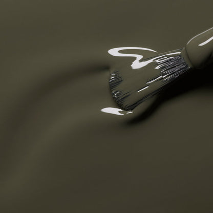 A close-up of a brush applying Manucurist's Khaki paint—a smooth, glossy olive green with grey undertones. Light reflects off the wet surface as the bristles blend this unique shade evenly.