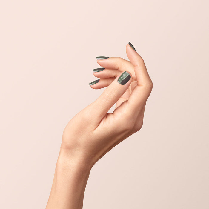 A hand with neatly manicured nails painted in Manucurist’s Khaki, an olive green polish with subtle grey undertones, is held gracefully against a pale, neutral background.