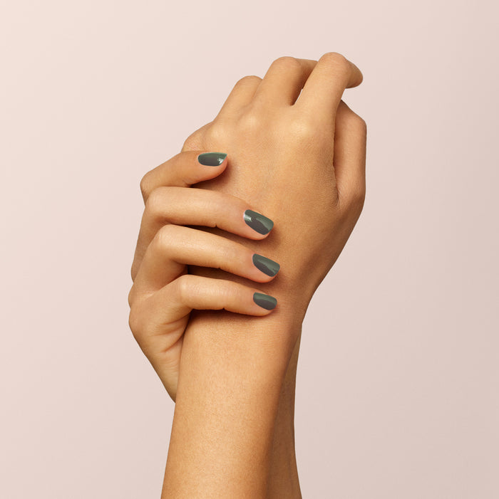 Two medium-toned hands rest on a light beige background. Their short nails are painted with Manucurist's Khaki, a matte gray polish with subtle grey undertones. One hand gently holds the other's wrist.