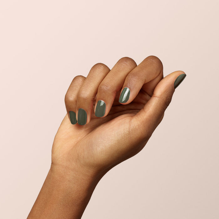 A hand with medium brown skin is shown against a beige background, nails neatly manicured and painted in Manucurist's Khaki—a Green Flash gel polish with subtle grey undertones. The hand is curled slightly inward, palm facing away.