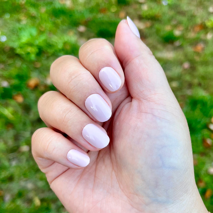 Kitten | Extra Short | Round - Tab Press-On showing soft pale neutral pink press-on nails on hand outdoors with natural green background