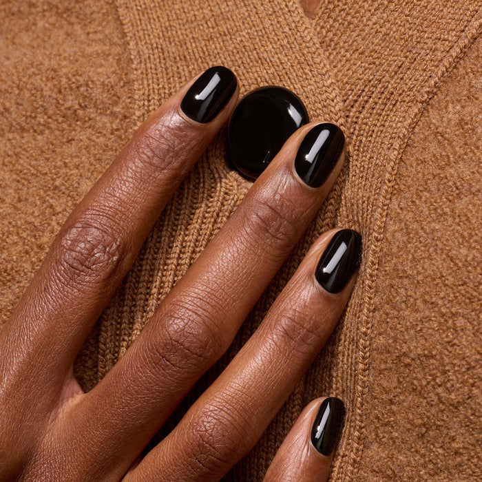 A hand with Manucurist's Licorice black nail polish rests on a brown wool sweater near a large black button.