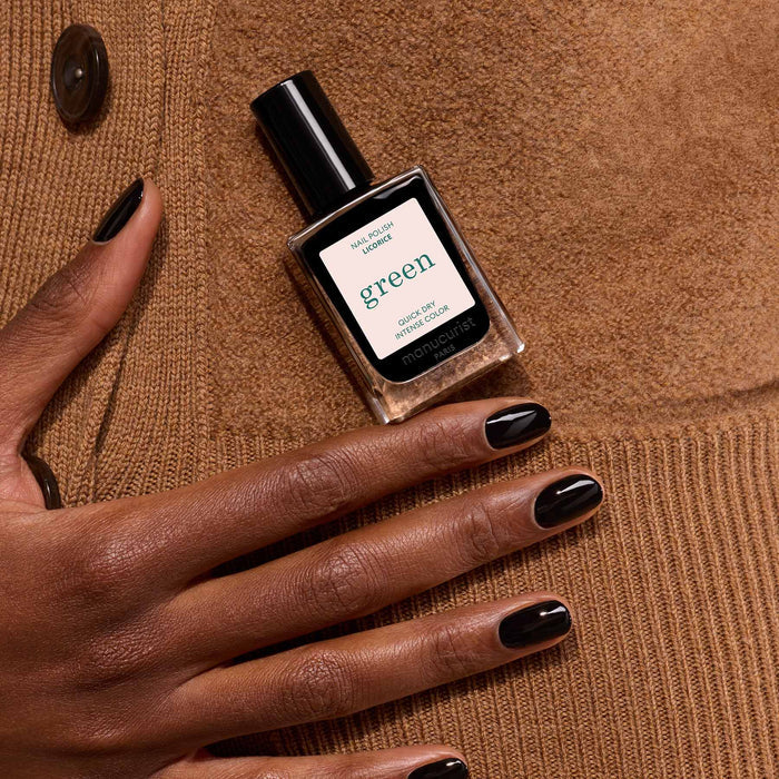 A hand with chic black, glossy polished nails rests on brown suede, holding a bottle of Manucurist Licorice nail color.